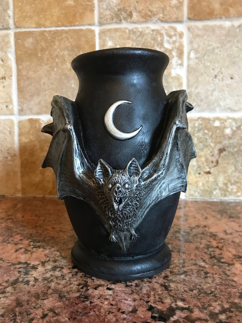 May include: Black ceramic vase with a bat and crescent moon design. The bat is silver and has a detailed design. The vase has a wide mouth and a rounded base.