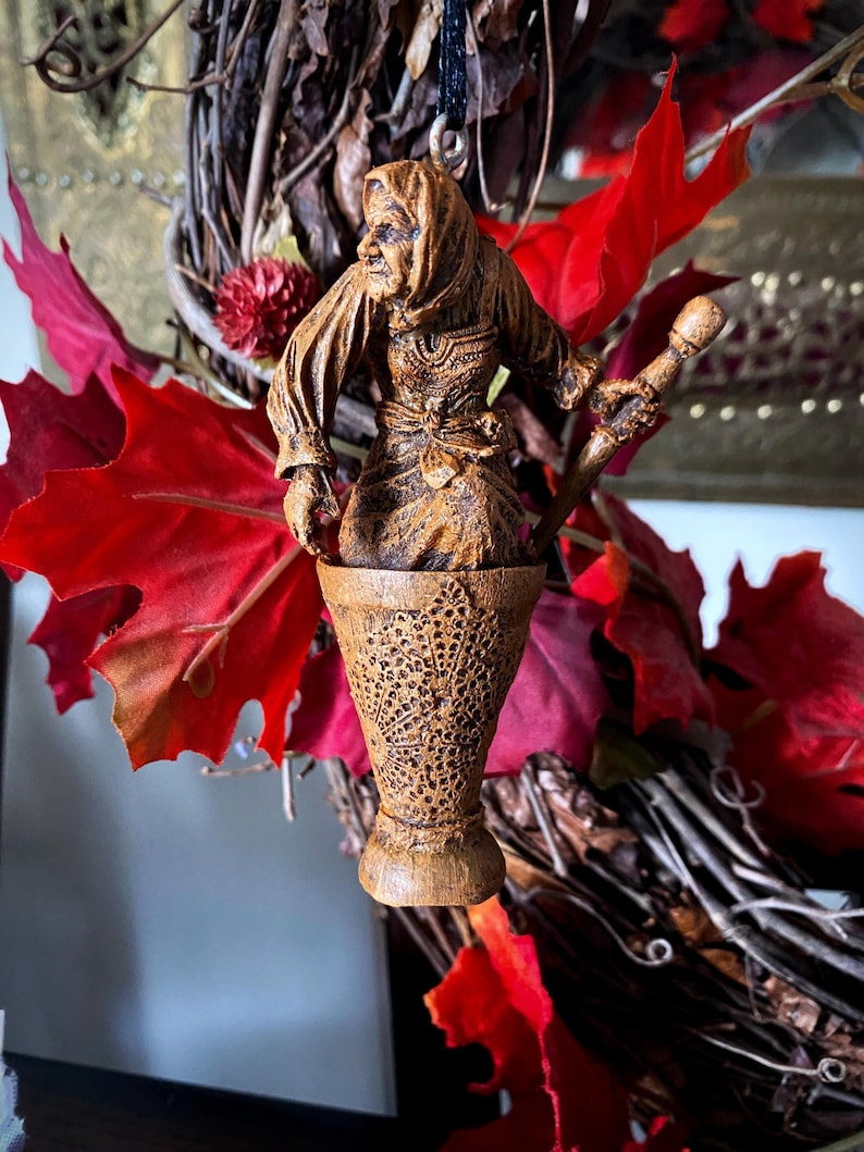 May include: A wooden figurine of a woman with a long, flowing robe and a staff. The figurine is carved in a detailed style and has a rustic, aged appearance. The figurine is hanging from a black cord and is suspended inside a wooden cup.