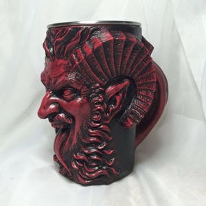 May include: A red ceramic mug with a detailed sculpted face of a demon with horns and a beard. The mug has a black and red color scheme.