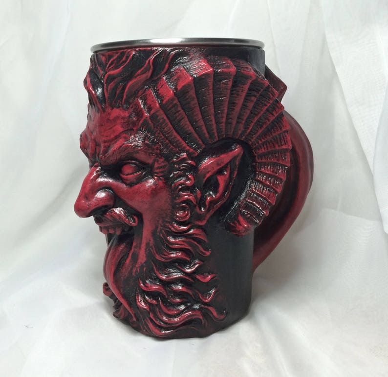 May include: A red ceramic mug with a detailed sculpted face of a demon with horns and a beard. The mug has a black and red color scheme.