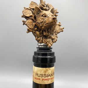Op de afbeelding: Een gouden flesstopper met een gedetailleerd gesculpteerd wolfshoofd, omringd door bladeren en takken. De stopper zit bovenop een wijnfles met een etiket waarop "RUSSIAN RIVER NOIR PINOT NOIR" staat.
