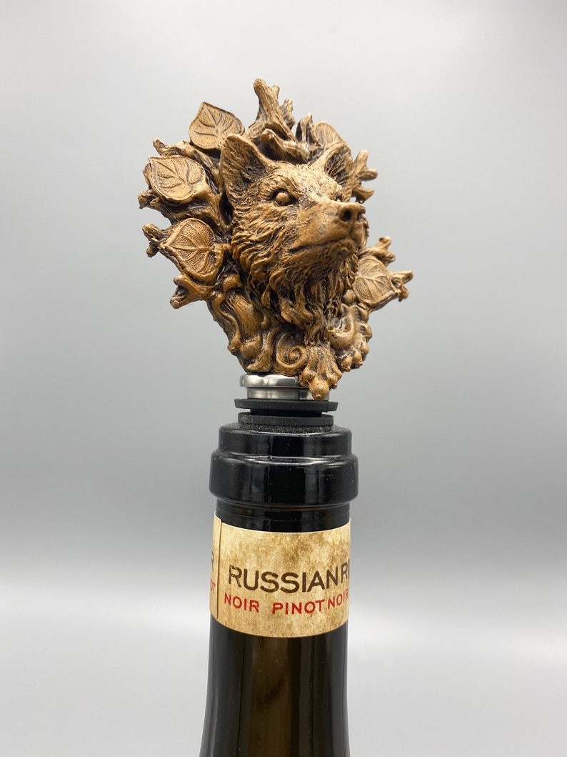 Op de afbeelding: Een gouden flesstopper met een gedetailleerd gesculpteerd wolfshoofd, omringd door bladeren en takken. De stopper zit bovenop een wijnfles met een etiket waarop "RUSSIAN RIVER NOIR PINOT NOIR" staat.