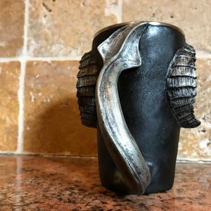 May include: Black ceramic mug with a silver handle shaped like a ram's horn. The mug has a textured surface and is decorated with silver ram's horn details.