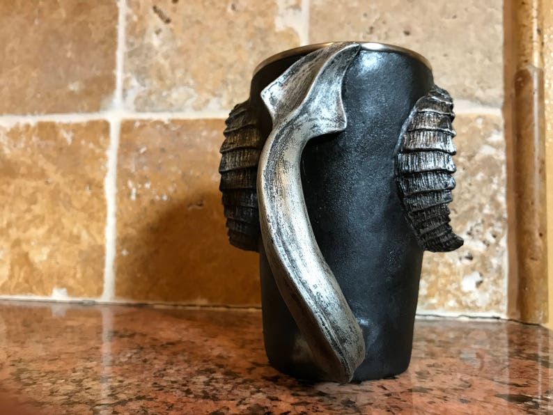 May include: Black ceramic mug with a silver handle shaped like a ram's horn. The mug has a textured surface and is decorated with silver ram's horn details.