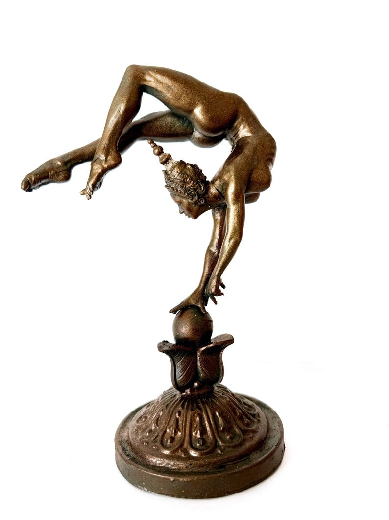 May include: A bronze sculpture of a woman performing a yoga pose, balancing on a ball atop a decorative base.