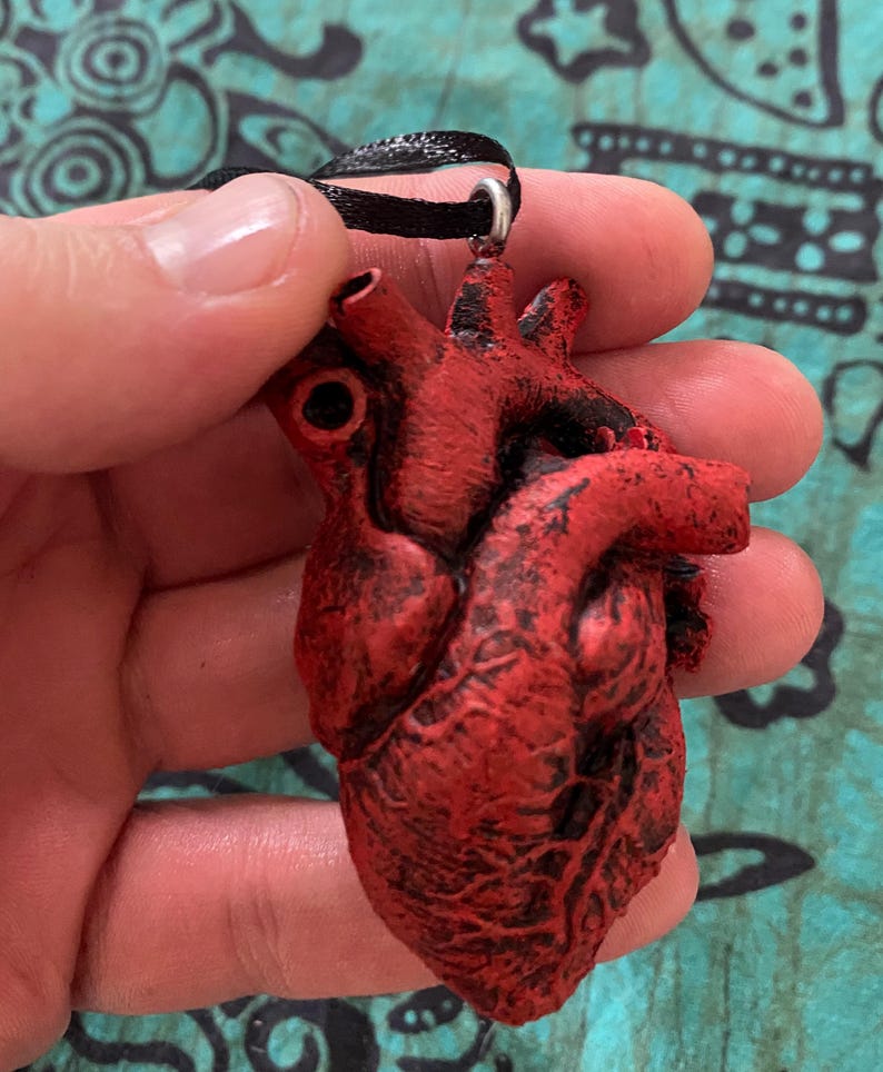 May include: A red anatomical heart pendant with a detailed texture, hanging from a black ribbon.
