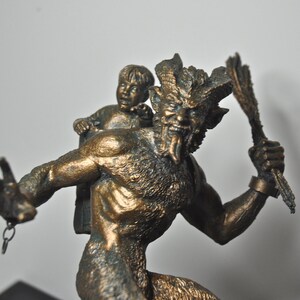 May include: A bronze sculpture of a horned, goat-like creature carrying a small child on its back. The creature has a fierce expression and is holding a staff in its hand.