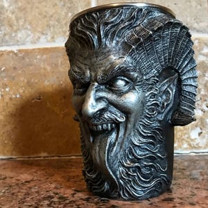 May include: A silver-colored metal mug shaped like a demon's head with a beard and horns. The mug has a detailed design and a textured surface.