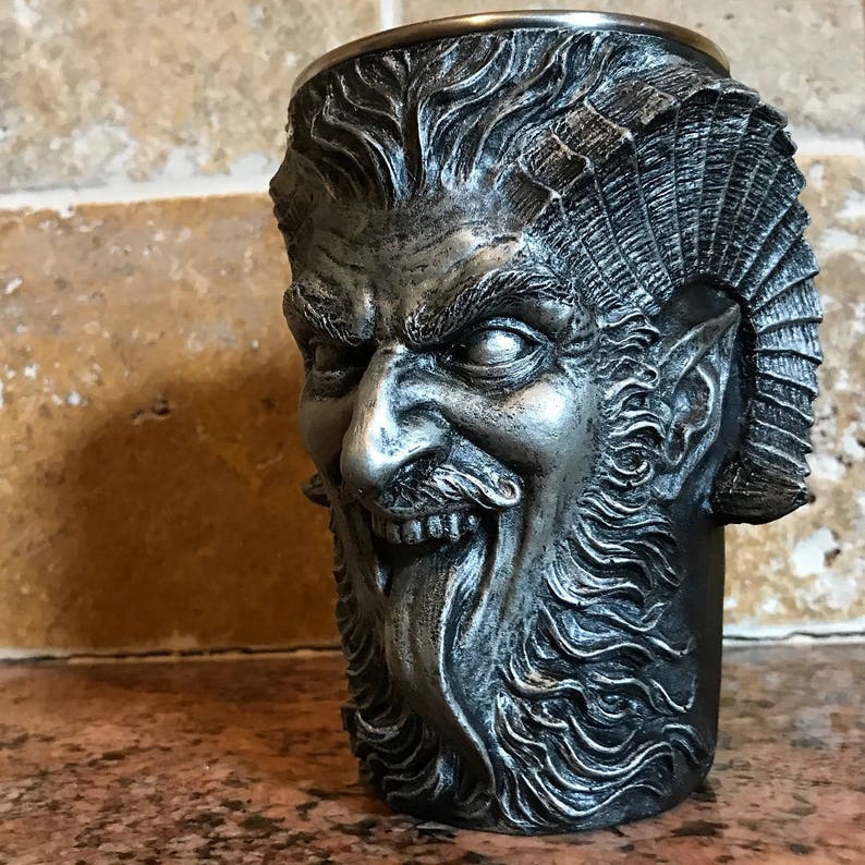 May include: A silver-colored metal mug shaped like a demon's head with a beard and horns. The mug has a detailed design and a textured surface.