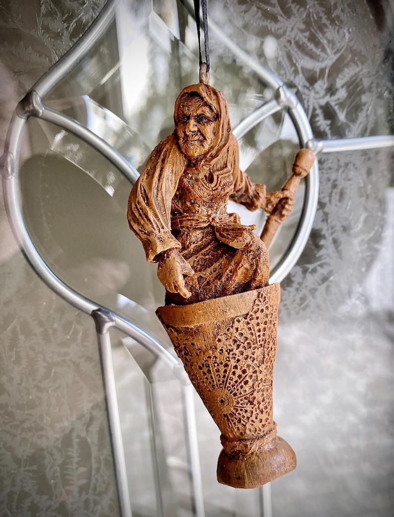 May include: A wooden figurine of a woman with a long white beard and a pointed hat, holding a staff. The figurine is carved in a rustic style and is painted in shades of brown. The figurine is hanging from a string.