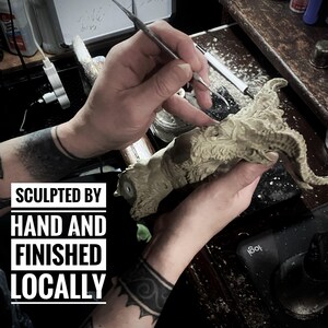 May include: A close-up of a person's hands sculpting a detailed, gray, clay sculpture of a creature with horns. The text "SCULPTED BY HAND AND FINISHED LOCALLY" is visible in the image.