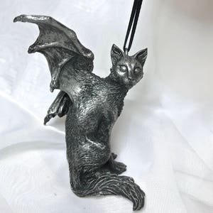 May include: A silver-colored figurine of a cat with bat wings. The cat is sitting with its head tilted to the side and its wings spread out behind it.