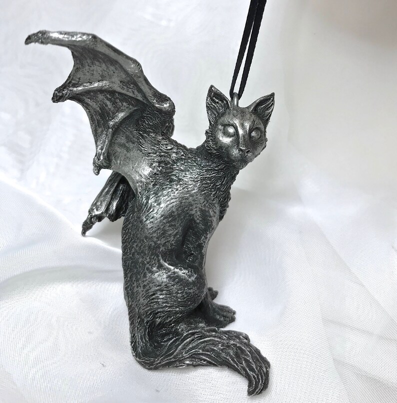 May include: A silver-colored figurine of a cat with bat wings. The cat is sitting with its head tilted to the side and its wings spread out behind it.