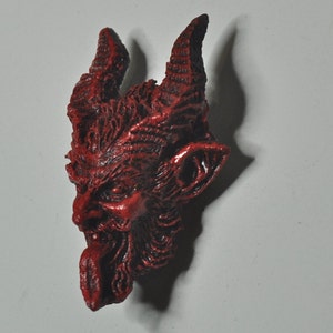May include: A red, detailed, demonic face with horns and a protruding tongue. The face is made of a textured material and has a slightly menacing expression.