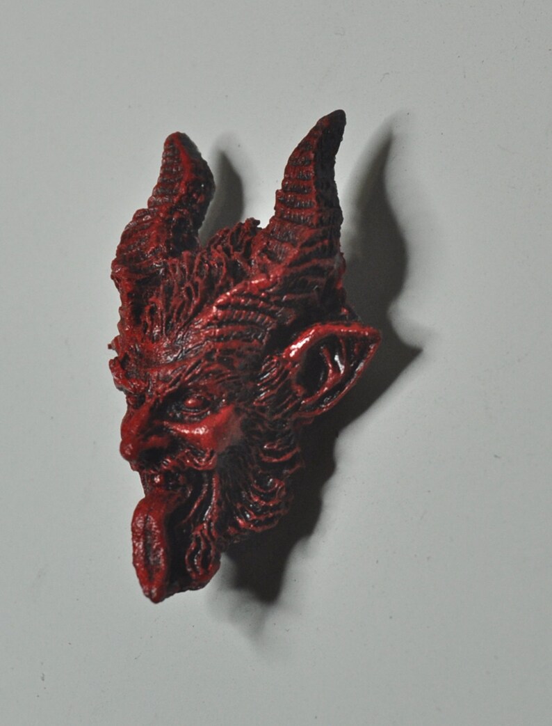 May include: A red, detailed, demonic face with horns and a protruding tongue. The face is made of a textured material and has a slightly menacing expression.