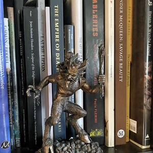 May include: A bronze-colored figurine of a horned creature with a tail, standing on a rocky base. The figurine is positioned on a bookshelf with a variety of books behind it.