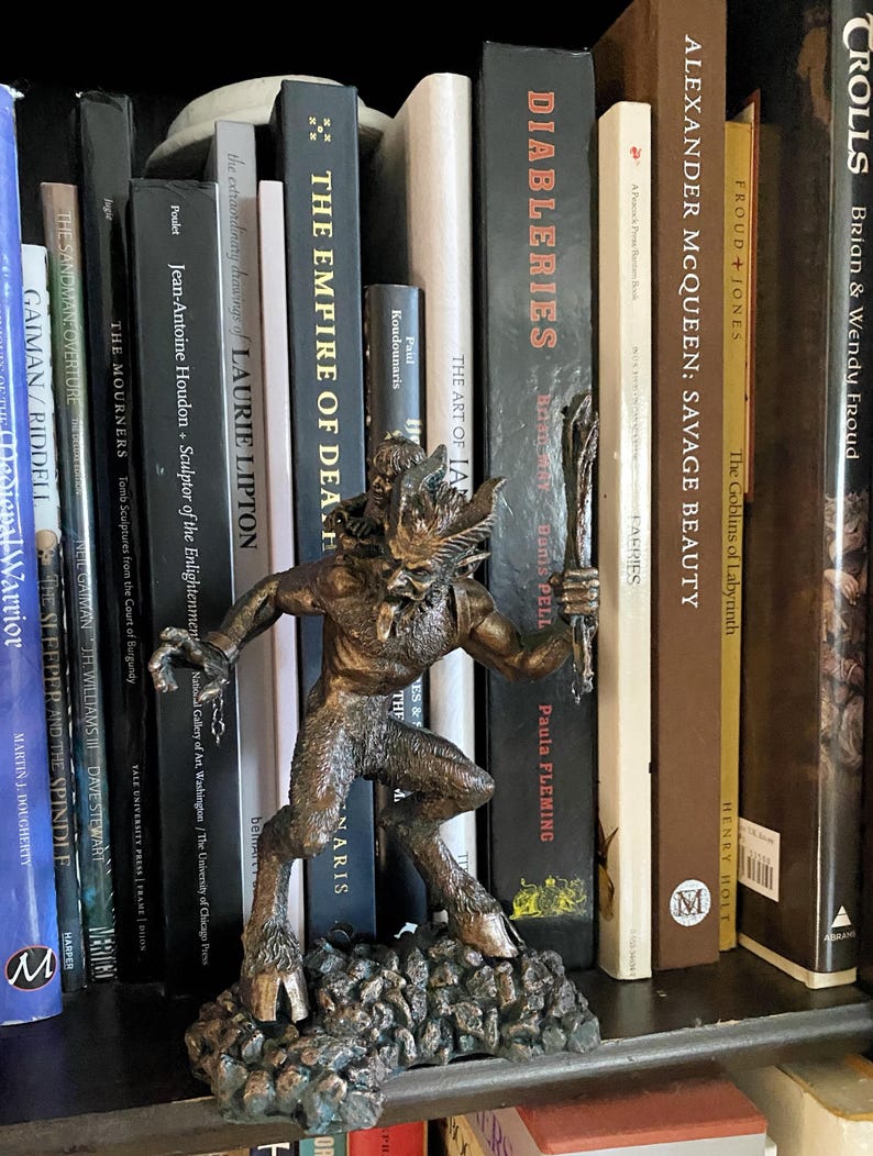 May include: A bronze-colored figurine of a horned creature with a tail, standing on a rocky base. The figurine is positioned on a bookshelf with a variety of books behind it.