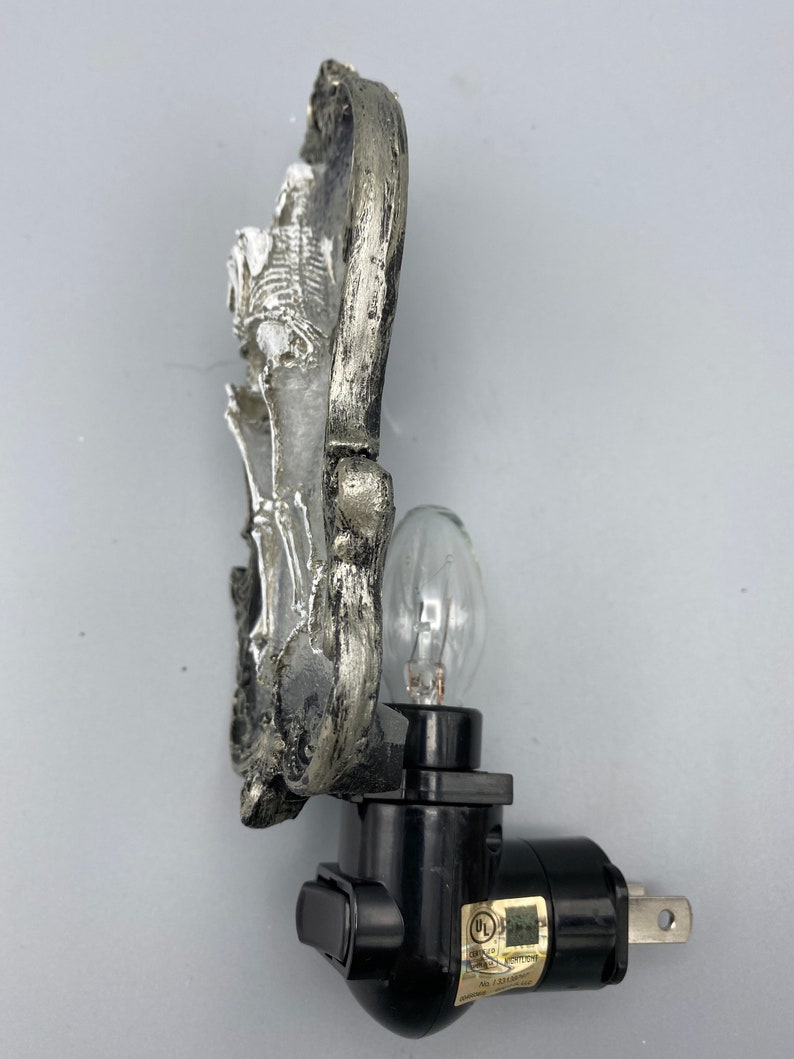 May include: A silver skeleton night light with a black plug-in base and an on/off switch. The light is shaped like a skull with a decorative design. The light is UL listed.