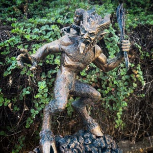 May include: A bronze statue of a horned, goat-like creature with a human-like body. The creature is holding a staff in its right hand and has a small human figure on its back. The statue is standing on a rocky base.