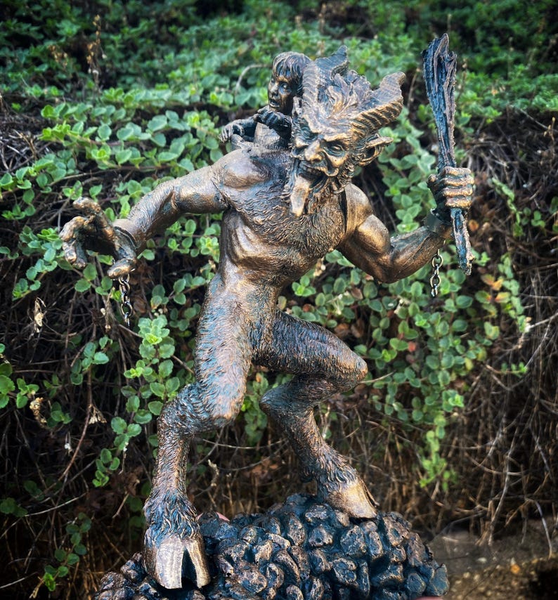 May include: A bronze statue of a horned, goat-like creature with a human-like body. The creature is holding a staff in its right hand and has a small human figure on its back. The statue is standing on a rocky base.