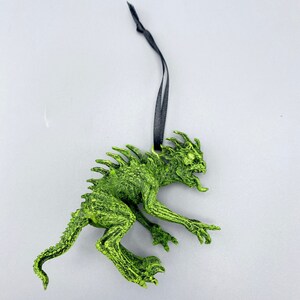 May include: Green monster ornament with a black ribbon for hanging. The monster has a spiky back, sharp teeth, and a long tail.