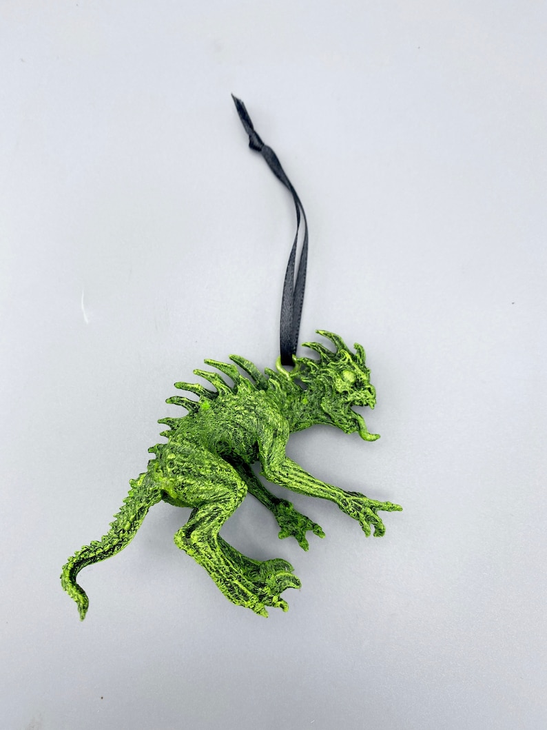 May include: Green monster ornament with a black ribbon for hanging. The monster has a spiky back, sharp teeth, and a long tail.