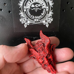 May include: A red resin sculpture of a demon's head with horns and a long tongue. The sculpture is detailed with textured lines and a menacing expression.