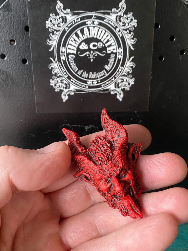 May include: A red resin sculpture of a demon's head with horns and a long tongue. The sculpture is detailed with textured lines and a menacing expression.
