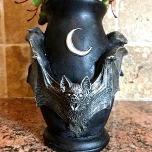 May include: Black ceramic vase with a bat design and a crescent moon on the front. The vase is decorated with silver paint and has a textured surface.