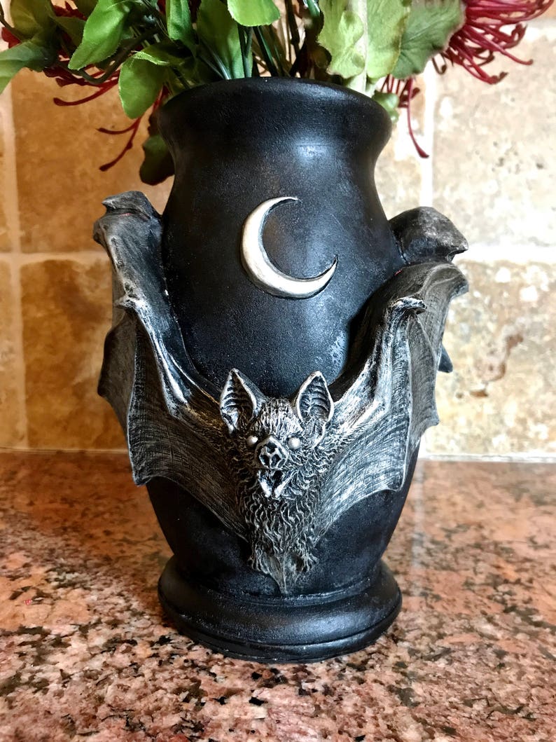 May include: Black ceramic vase with a bat design and a crescent moon on the front. The vase is decorated with silver paint and has a textured surface.