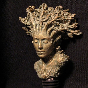 May include: A bronze bottle stopper featuring a woman's face with a crown of leaves. The stopper is made of metal and has a black rubber seal.