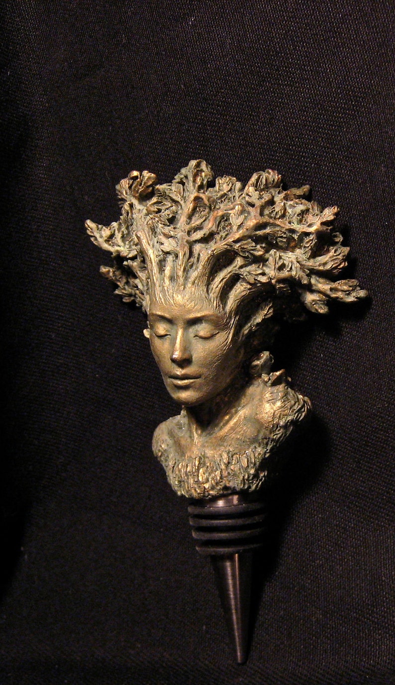 May include: A bronze bottle stopper featuring a woman's face with a crown of leaves. The stopper is made of metal and has a black rubber seal.