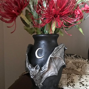 May include: Black ceramic vase with a bat and crescent moon design. The vase is filled with red and green artificial flowers.