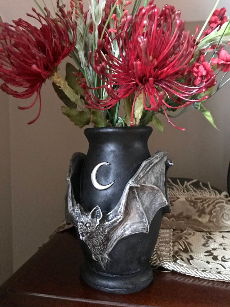May include: Black ceramic vase with a bat and crescent moon design. The vase is filled with red and green artificial flowers.