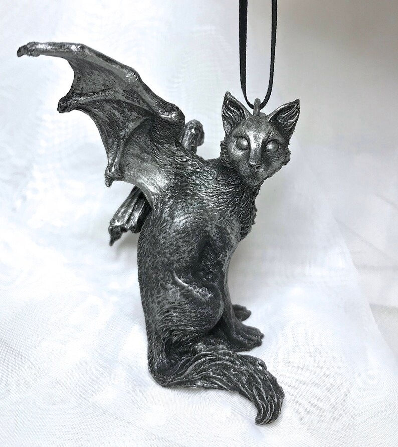 May include: A silver-colored winged cat figurine with a black cord attached to the top of its head. The cat is sitting with its wings spread out behind it.
