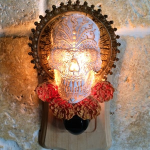 May include: A white sugar skull nightlight with intricate floral designs, surrounded by a gold-colored halo and red flowers. The skull is illuminated from within, creating a warm glow.