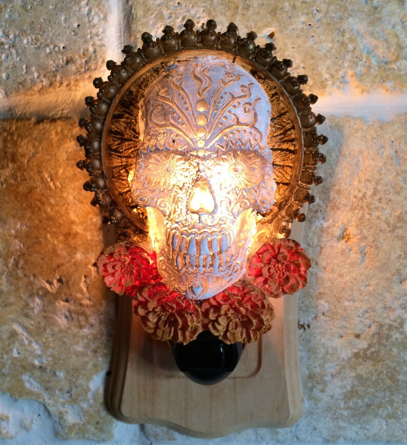 May include: A white sugar skull nightlight with intricate floral designs, surrounded by a gold-colored halo and red flowers. The skull is illuminated from within, creating a warm glow.
