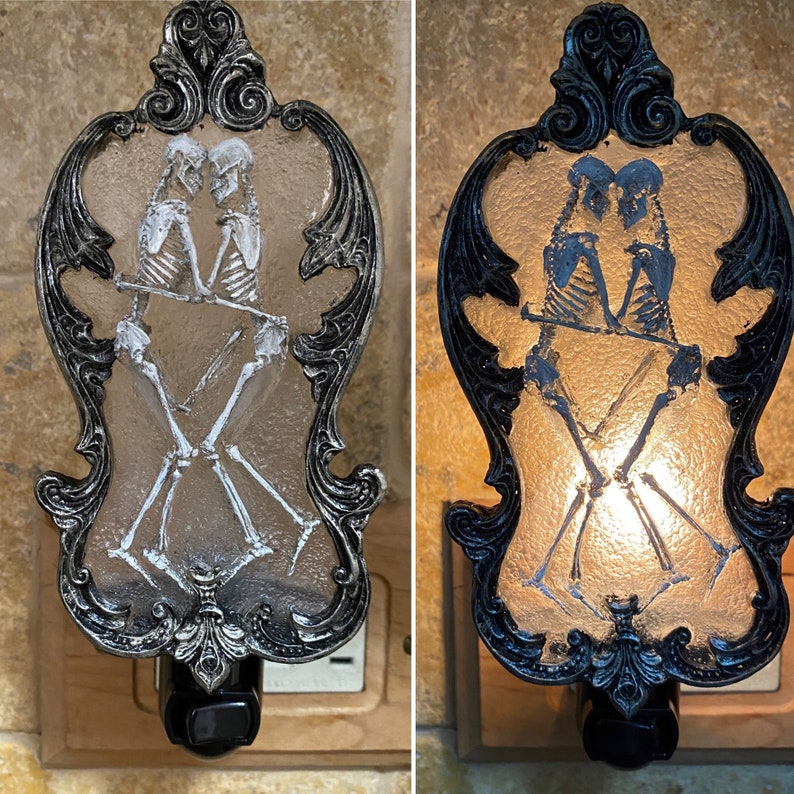 May include: A pair of skeletal figures in a romantic embrace are depicted on a black and silver nightlight. The nightlight is shaped like a decorative frame with ornate details.