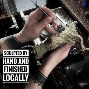 May include: A close-up of a person's hands sculpting a detailed, gray, clay sculpture of a creature with horns. The text "SCULPTED BY HAND AND FINISHED LOCALLY" is visible in the image.