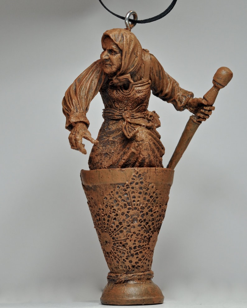 May include: A brown ceramic figurine of a woman holding a mortar and pestle. The woman is wearing a long brown dress and a brown shawl. The figurine is hanging from a black cord.