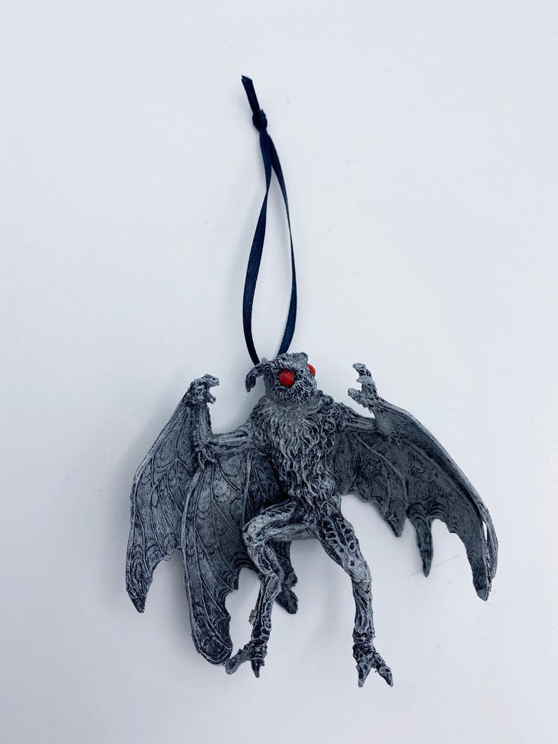 Puede incluir: Una criatura gris y blanca con forma de murci&eacute;lago con ojos rojos que cuelga de una cuerda negra. La criatura tiene alas grandes y garras afiladas. Es un adorno navide&ntilde;o.