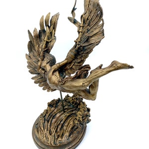 May include: A gold-toned sculpture of an angel with wings outstretched, falling into a wave. The angel is depicted with a flowing gown and a crown of thorns. The sculpture is mounted on a circular base.