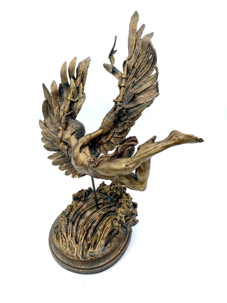 May include: A gold-toned sculpture of an angel with wings outstretched, falling into a wave. The angel is depicted with a flowing gown and a crown of thorns. The sculpture is mounted on a circular base.