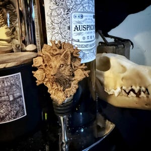Op de afbeelding: Een houten flessenstop met een gedetailleerde carving van een wolfskop omringd door bladeren. De stop ligt op een zwart oppervlak naast een fles wijn met een etiket waarop "AUSPICIOUS CABERNET SAUVIGNON" staat.