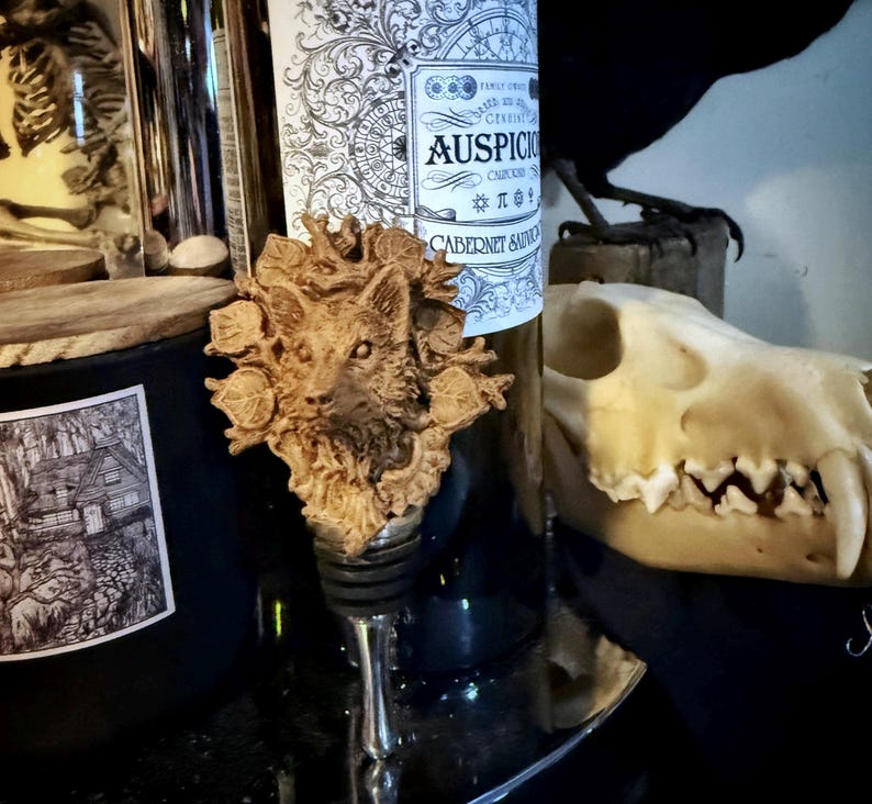 Op de afbeelding: Een houten flessenstop met een gedetailleerde carving van een wolfskop omringd door bladeren. De stop ligt op een zwart oppervlak naast een fles wijn met een etiket waarop "AUSPICIOUS CABERNET SAUVIGNON" staat.