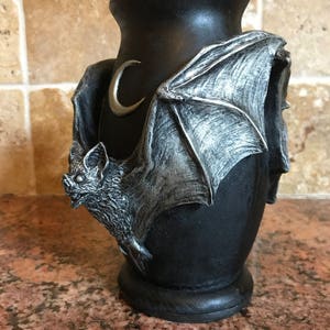 May include: Black ceramic vase with a silver bat and crescent moon design. The bat is sculpted with wings outstretched and a detailed face.