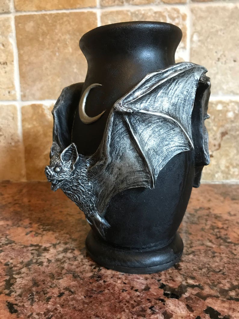 May include: Black ceramic vase with a silver bat and crescent moon design. The bat is sculpted with wings outstretched and a detailed face.