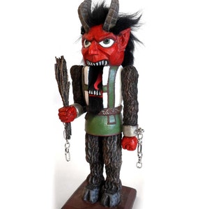 May include: A red and black wooden nutcracker figurine of a devil with horns, a green and white vest, and chains on his wrists. The nutcracker is holding a wooden stick in his right hand.