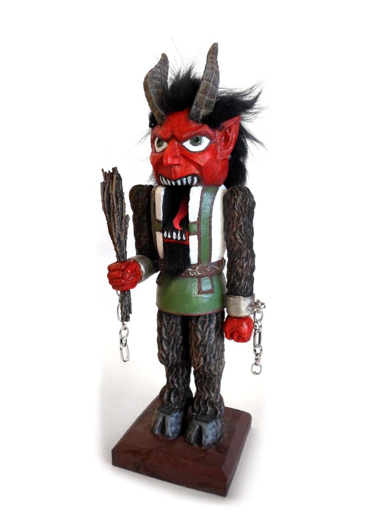 May include: A red and black wooden nutcracker figurine of a devil with horns, a green and white vest, and chains on his wrists. The nutcracker is holding a wooden stick in his right hand.