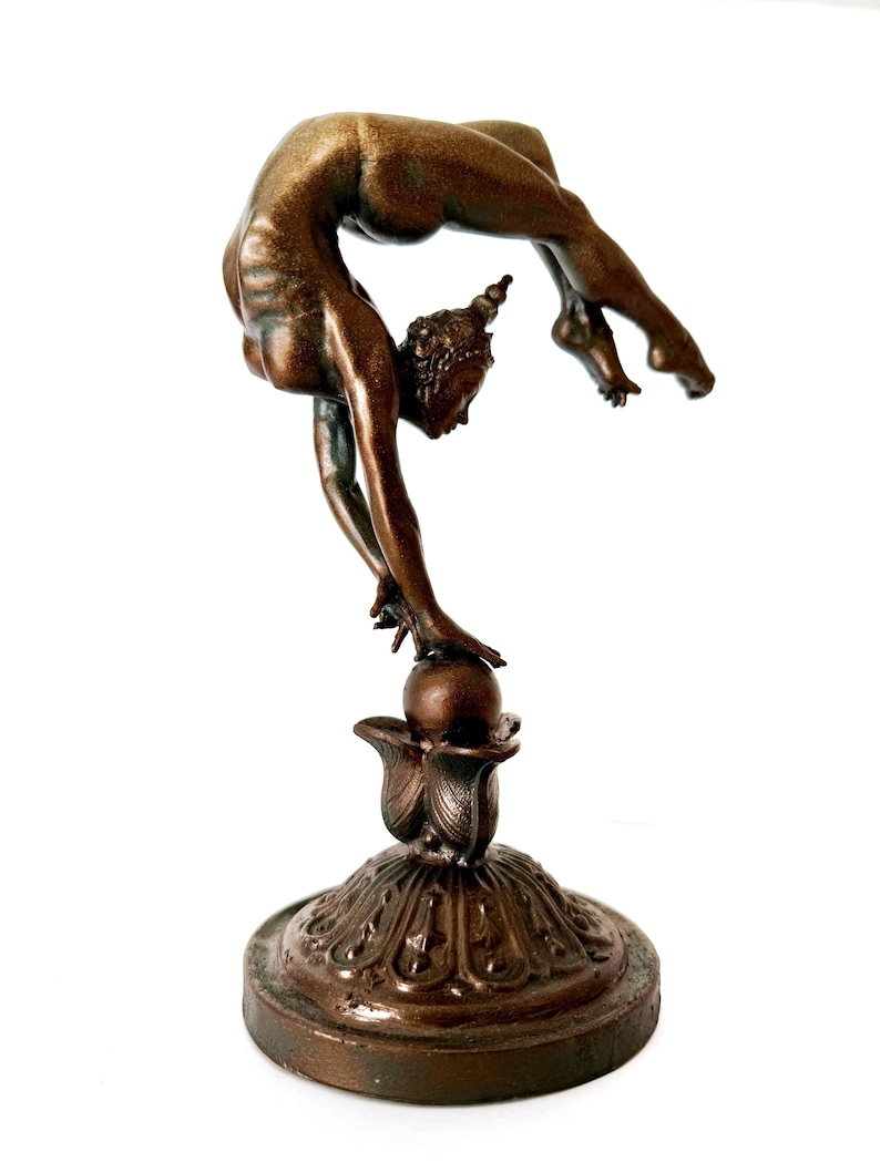 May include: A bronze sculpture of a woman performing a contortionist pose, balancing on a ball atop a decorative base.